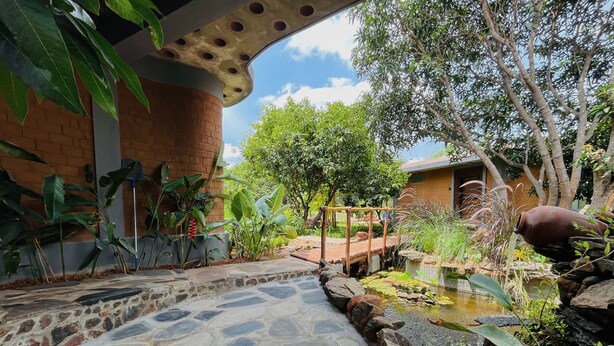 A stone pathway leading to a pond with a wooden bridge, surrounded by lush greenery and a brick building. - Dyavaaprthivi (Shoolagiri)