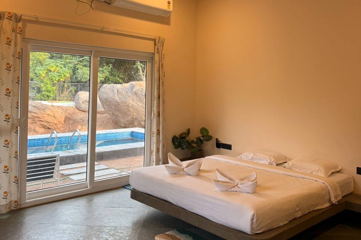 Habitación de hotel con cama, ventilador de techo, unidad de aire acondicionado, ventana con vista a una piscina y una planta pequeña.
