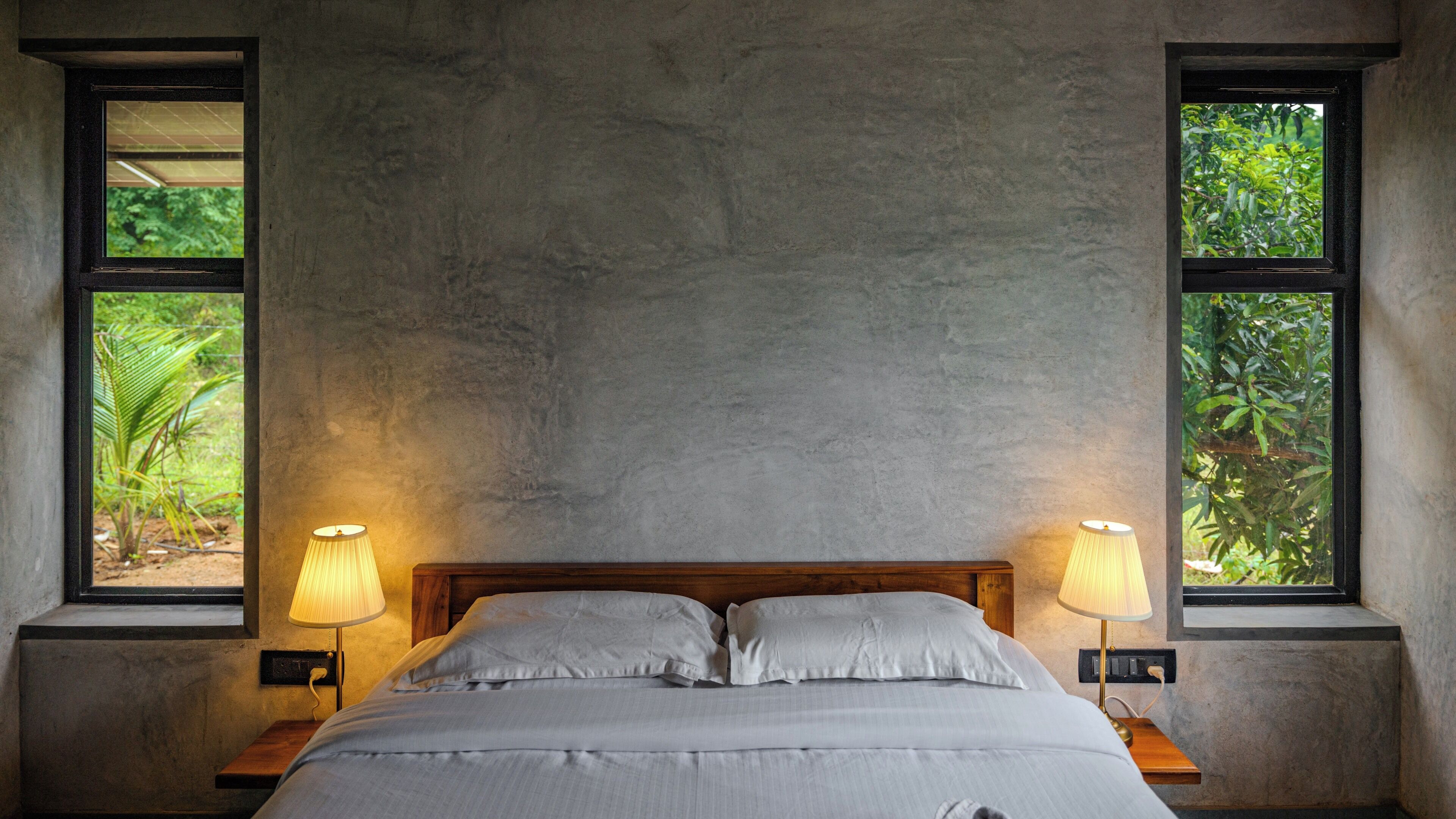 A bedroom with a bed, two bedside lamps, and two windows with views of greenery.