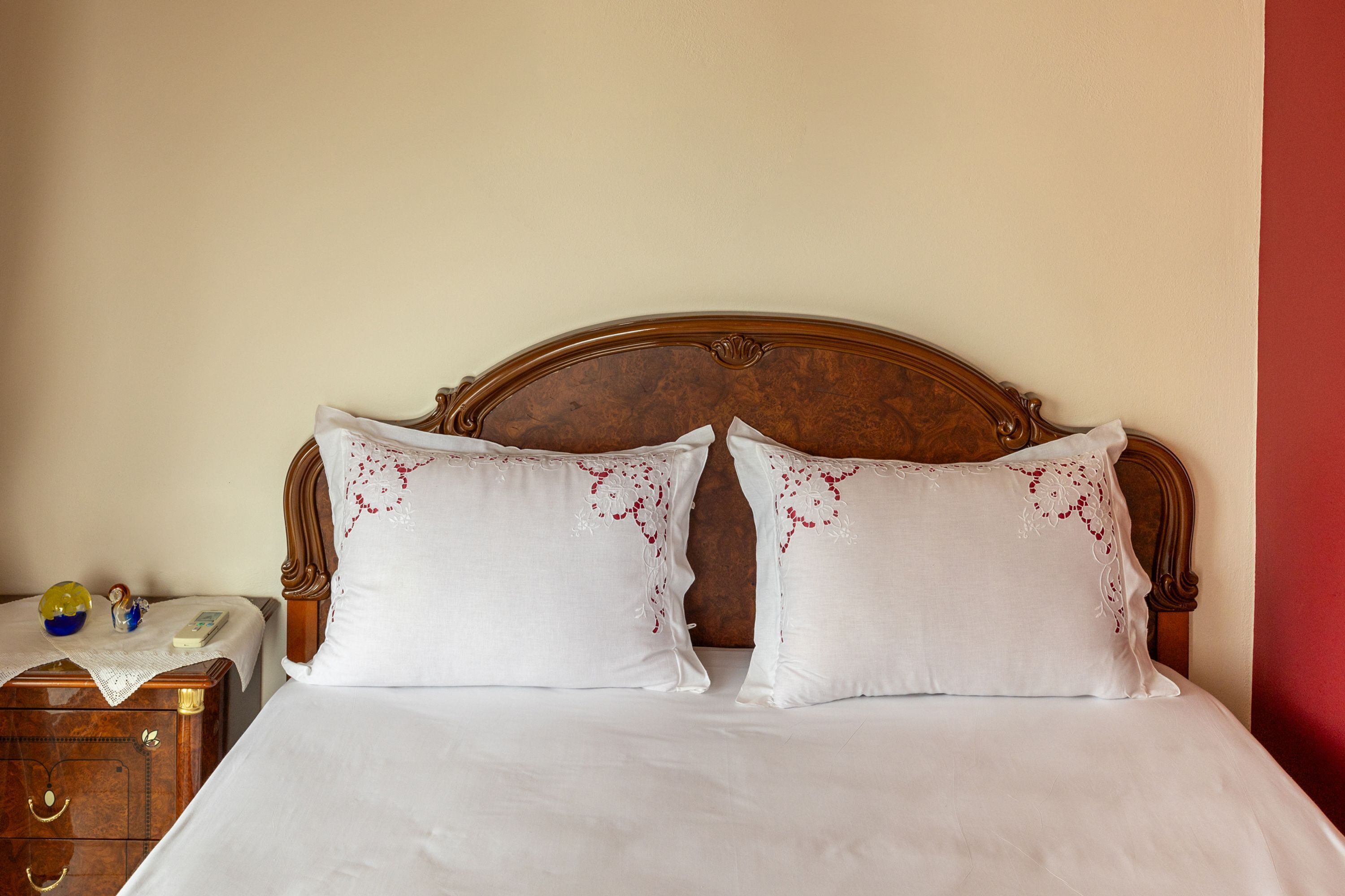 A neatly made bed with white linens and decorative pillows, a wooden nightstand with a telephone and a small figurine, and a plain wall with a red accent.