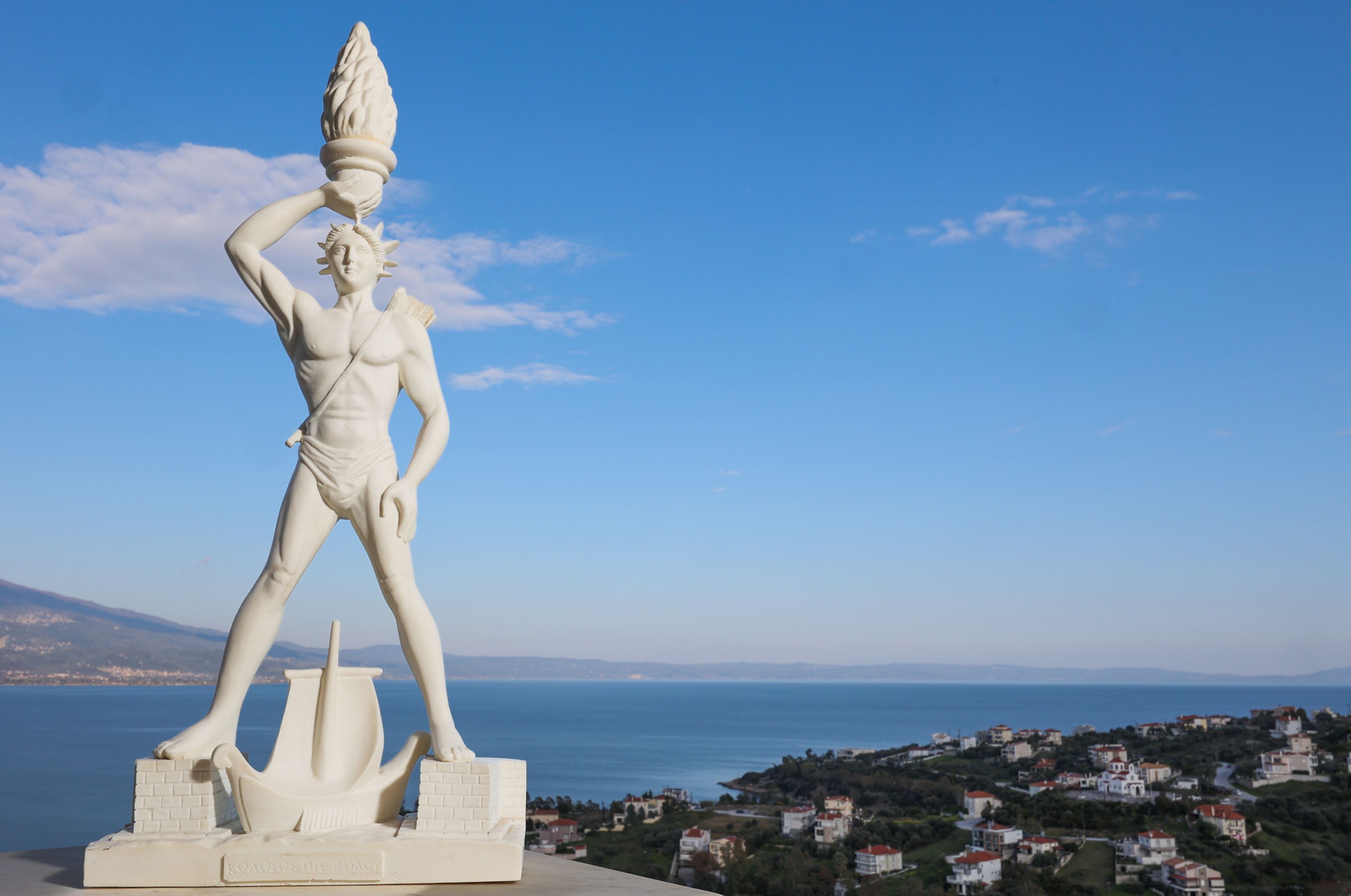 Una estatua de figura clásica sobre un pedestal, con un pueblo costero al fondo.