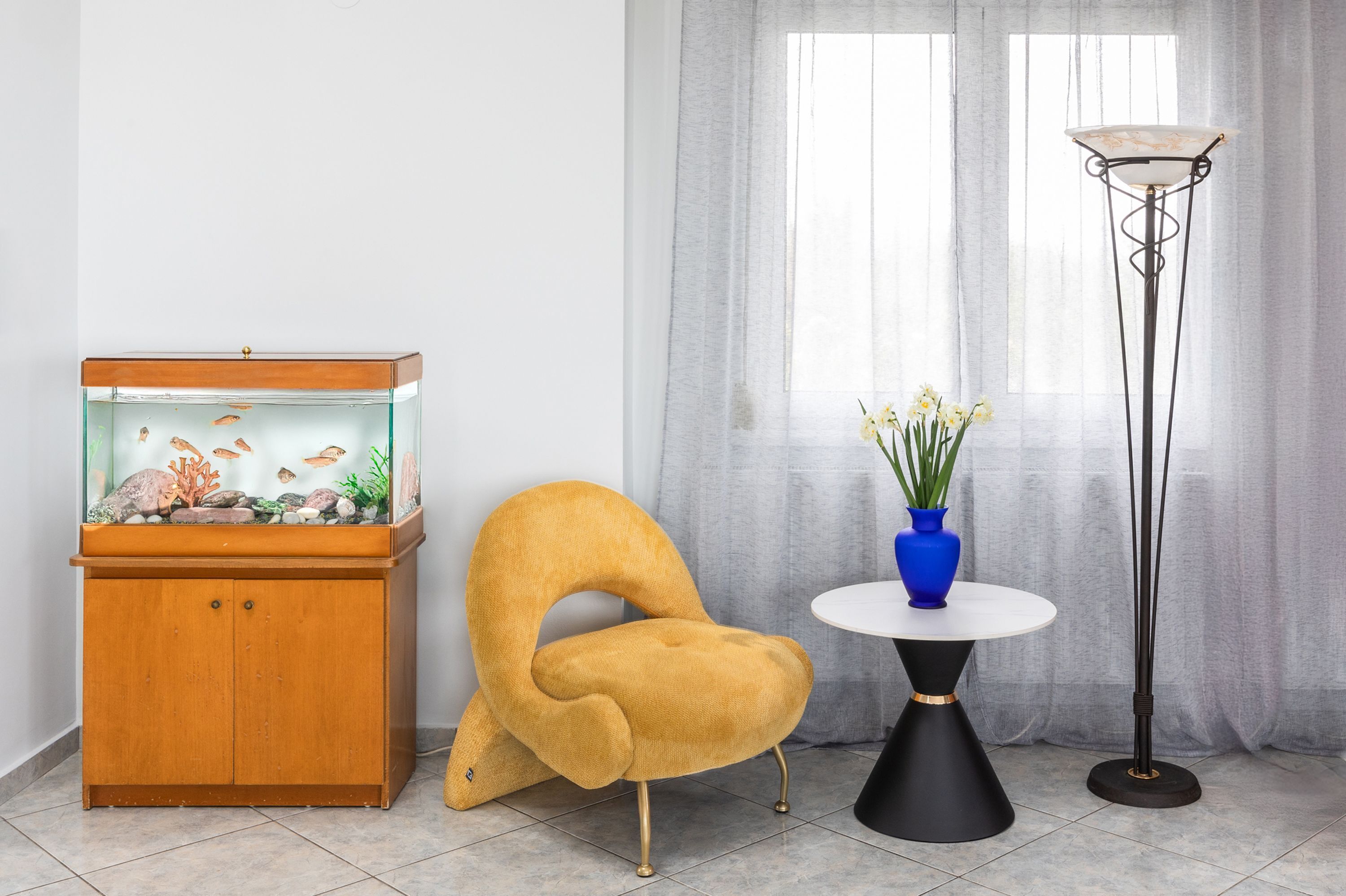 Een kamer met een vissenkom, een stoel, een tafel met een vaas en een vloerlamp.