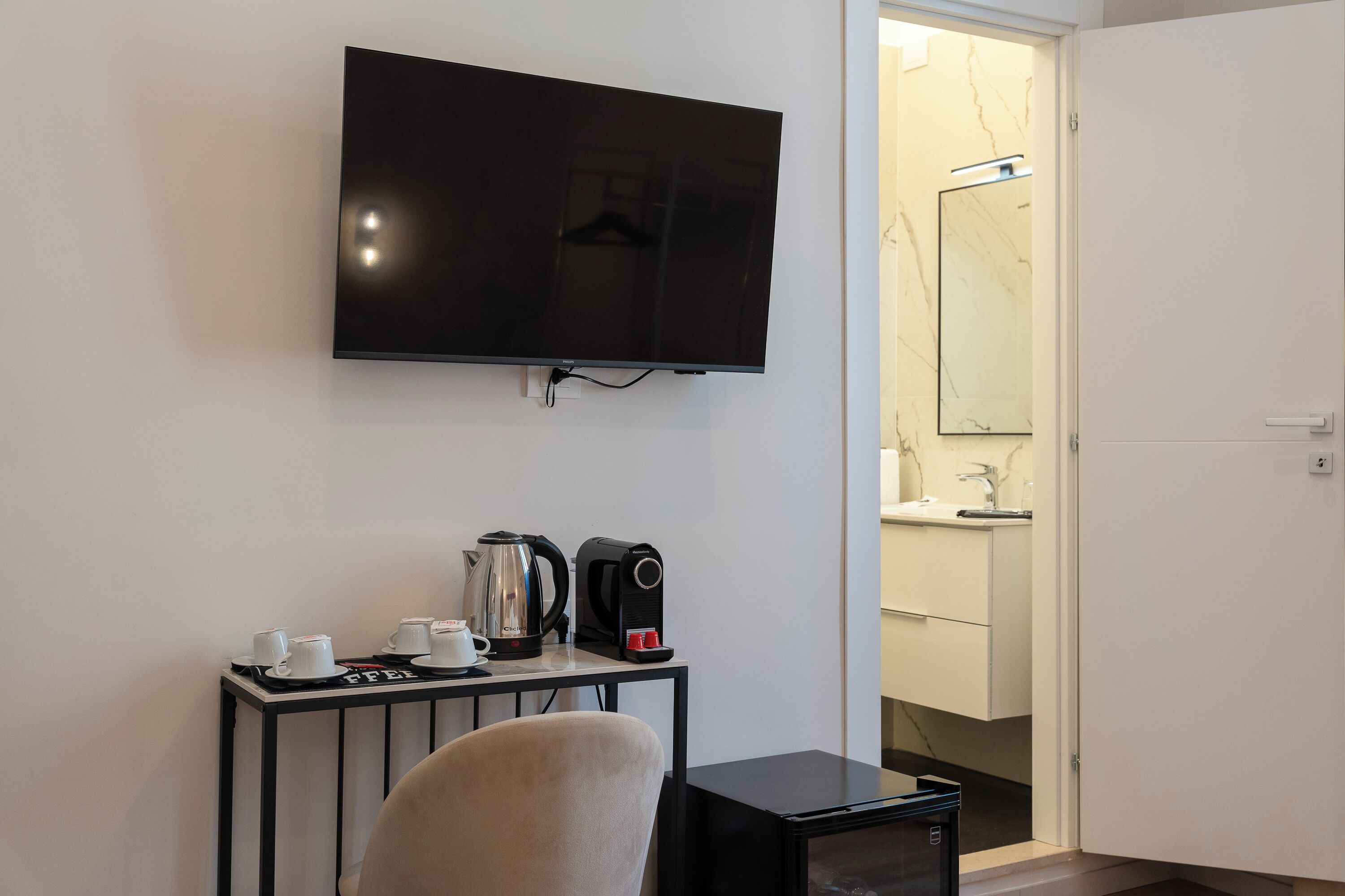 A hotel room with a flat-screen TV mounted on the wall, a small table with a kettle, cups, and a toaster, and a door leading to a bathroom.