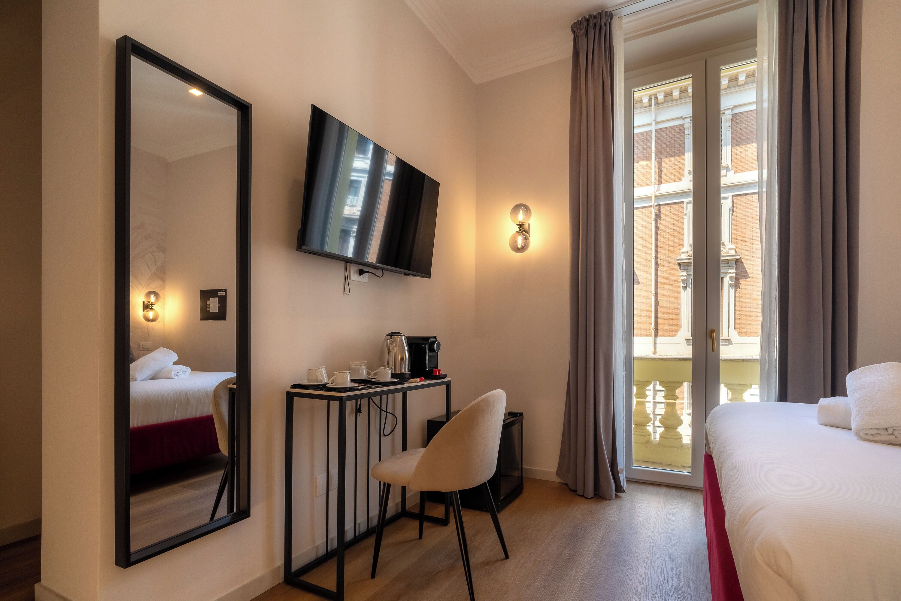 A hotel room with a bed, a desk with a coffee maker, a chair, a TV mounted on the wall, and a large window with curtains.
