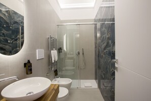Un baño moderno con un lavamanos ovalado grande, inodoro, ducha con mampara de vidrio y pared de mármol.