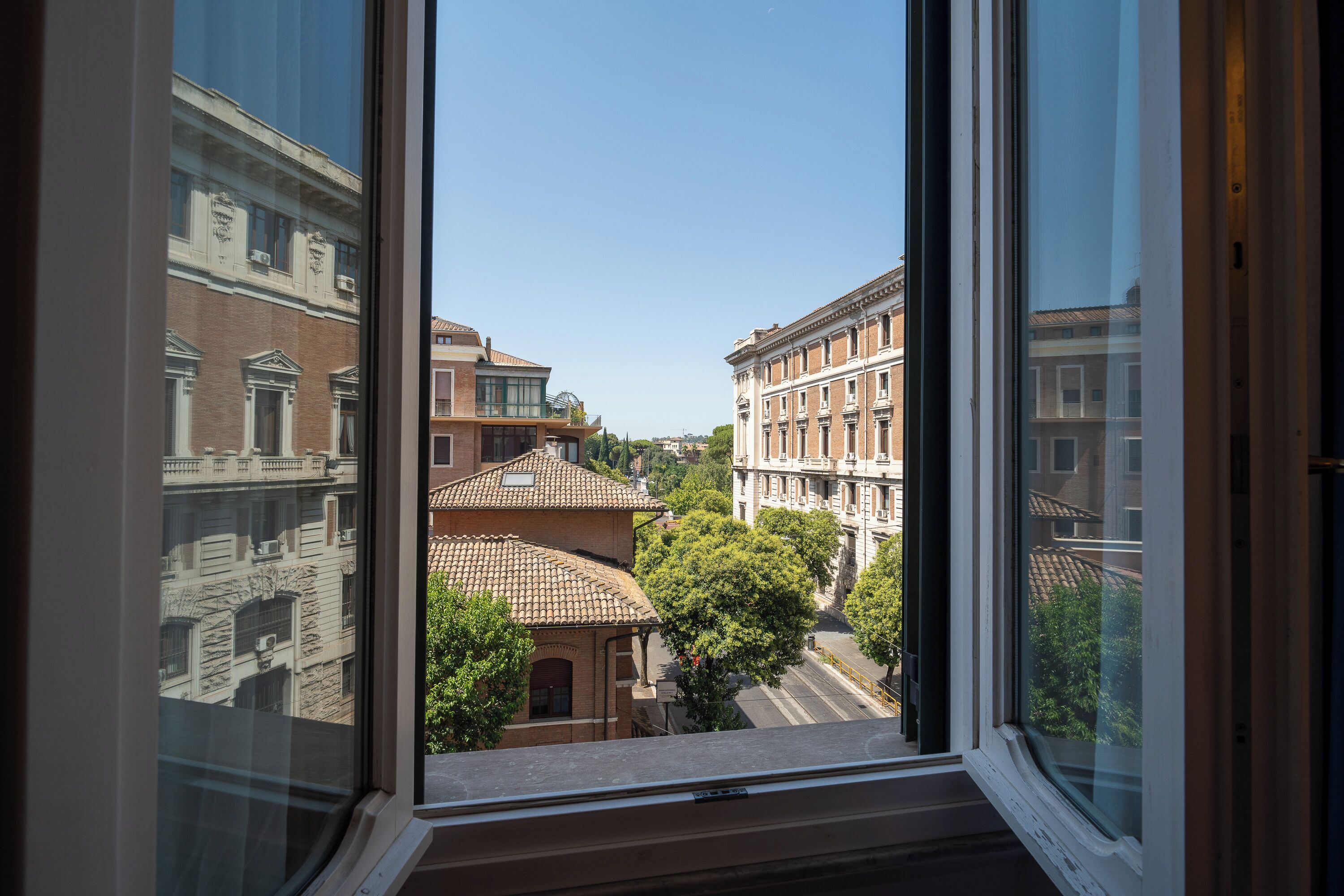 Vista a través de una ventana mostrando una calle con edificios, árboles y un cielo despejado.