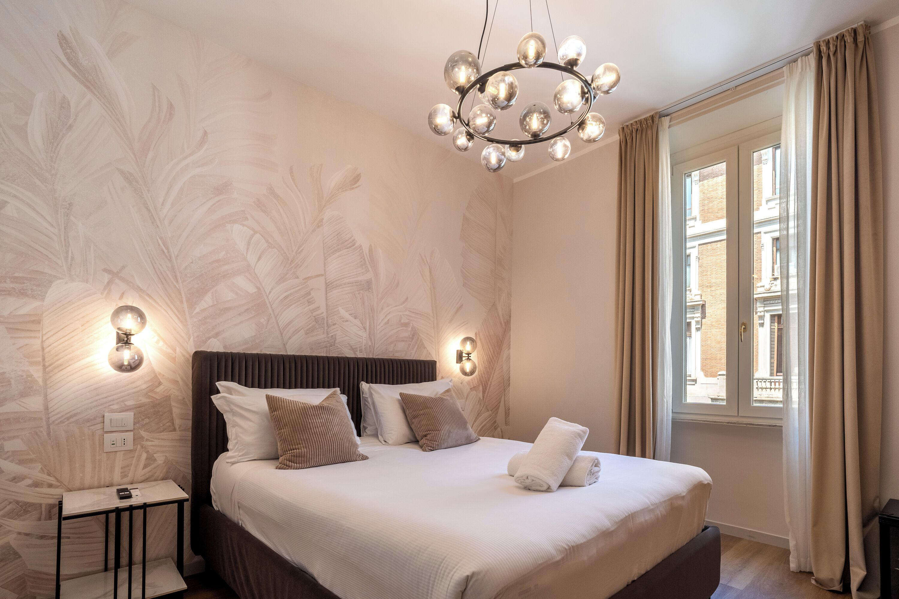Un dormitorio con cama, mesita de noche, lámpara de pared y ventana con cortinas.