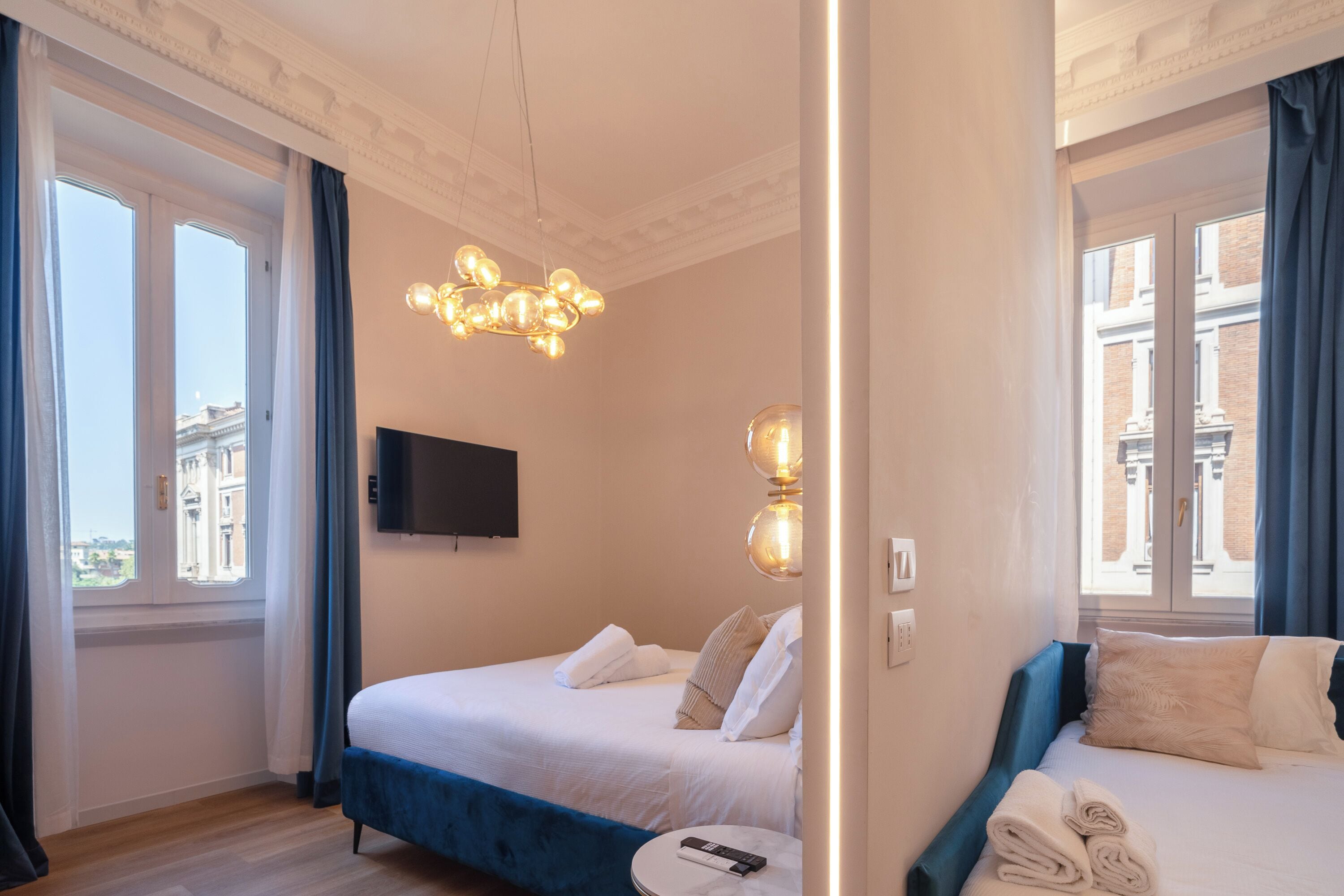 A hotel room with two beds, a flat-screen TV, a modern chandelier, and a view of buildings through large windows.