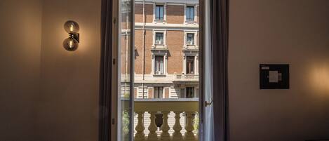 A hotel room with a bed, a window with curtains, a wall-mounted light fixture, and a view of a building with balconies.