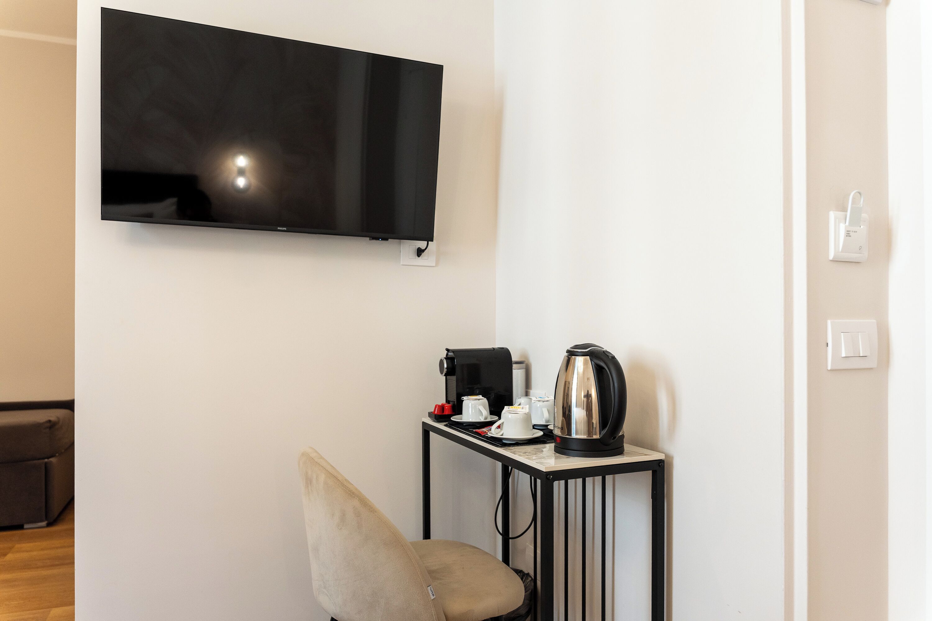 A room with a flat-screen TV mounted on the wall, a small table with a coffee maker, cups, and a kettle, and a single chair.