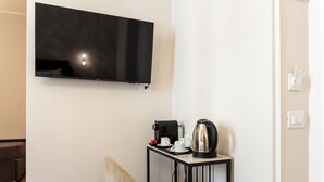 Una stanza con una televisione a schermo piatto montata a parete, un tavolino con una macchina del caffè, tazze e un bollitore, e una sedia.