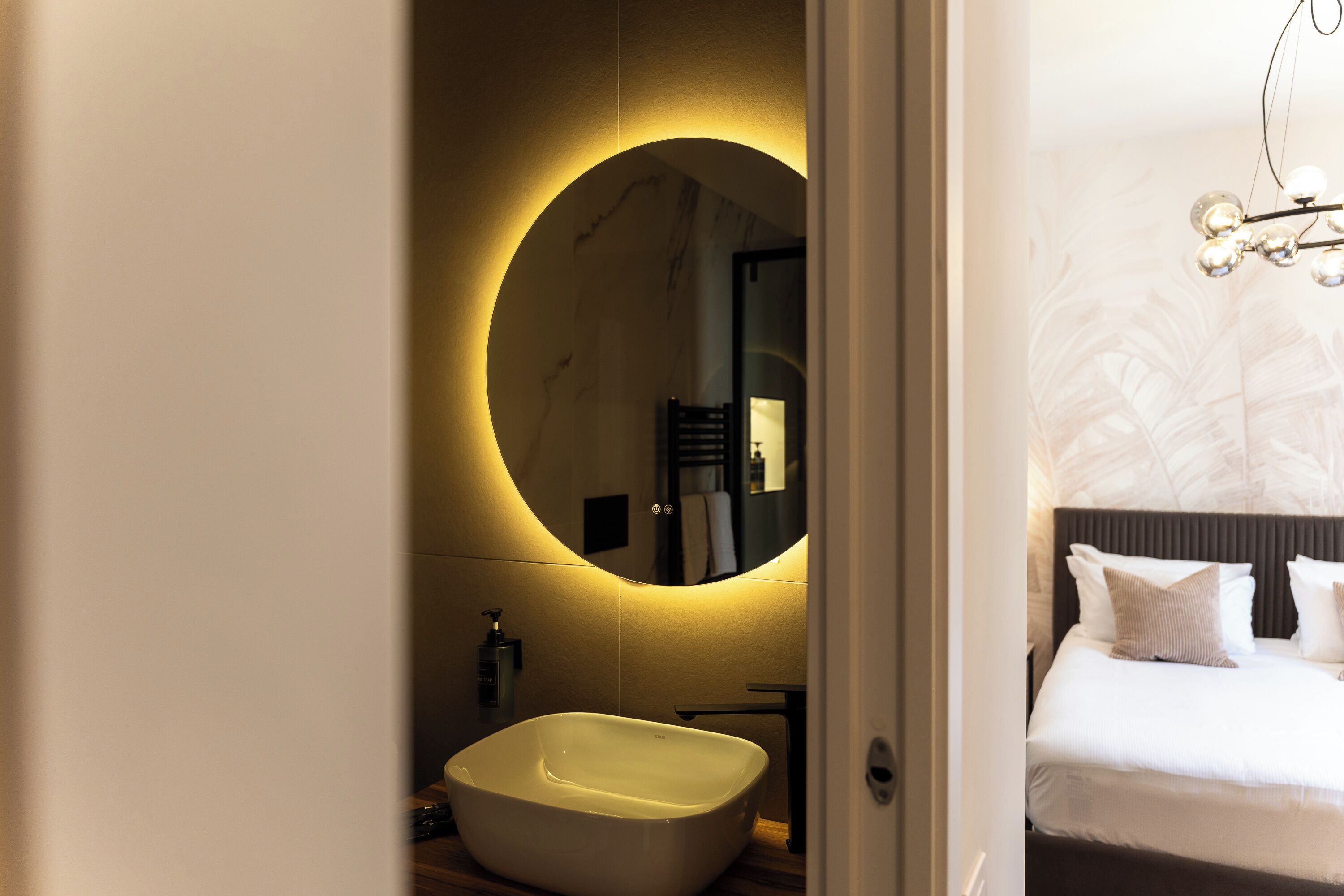 Une salle de bain moderne avec un miroir rond, un lavabo blanc et un lit avec une literie blanche.