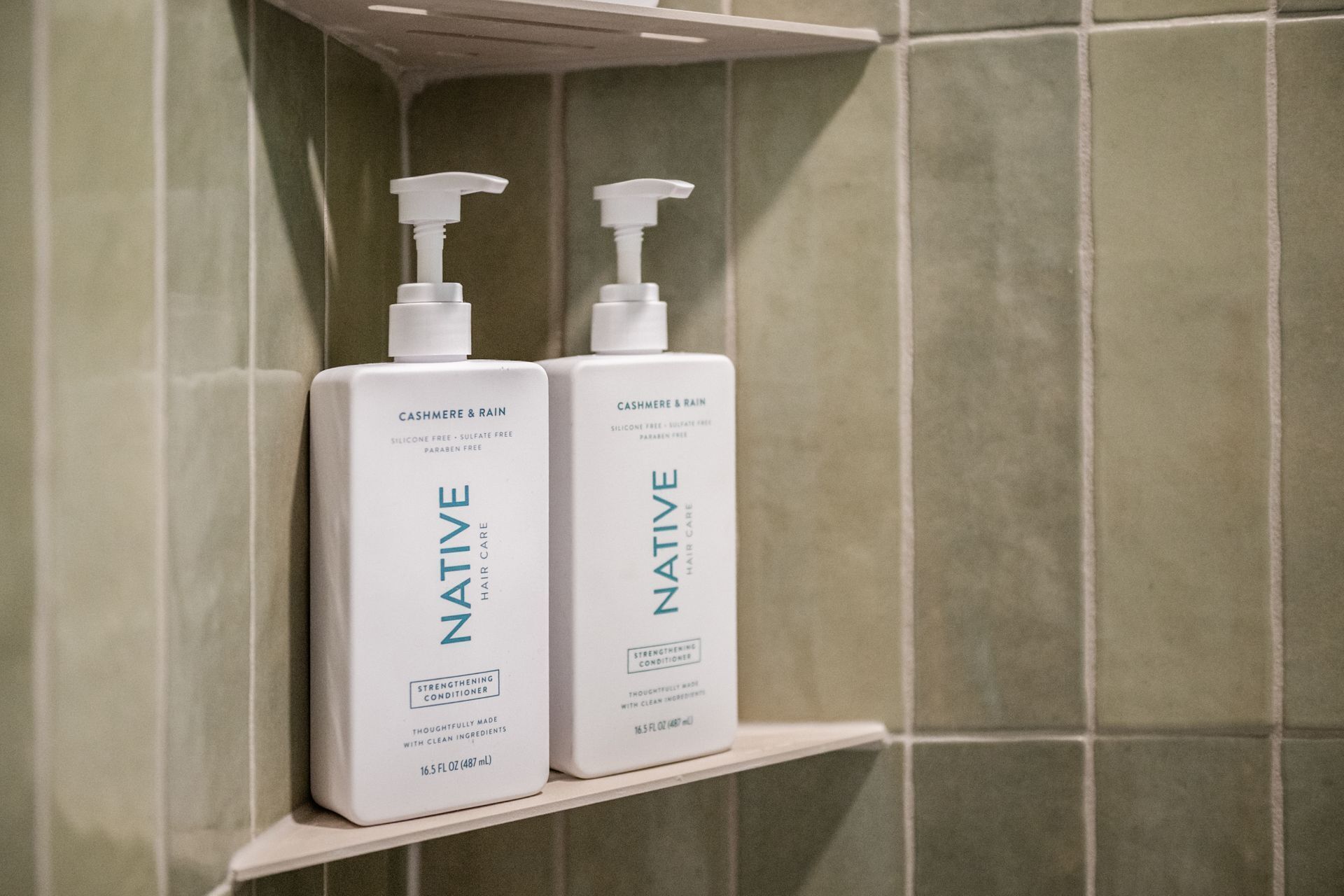 Dos botellas de acondicionador para el cabello Native sobre un estante blanco frente a una pared de azulejos.