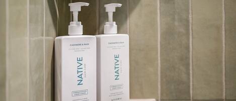 Dos botellas de acondicionador para el cabello Native sobre un estante blanco frente a una pared de azulejos.