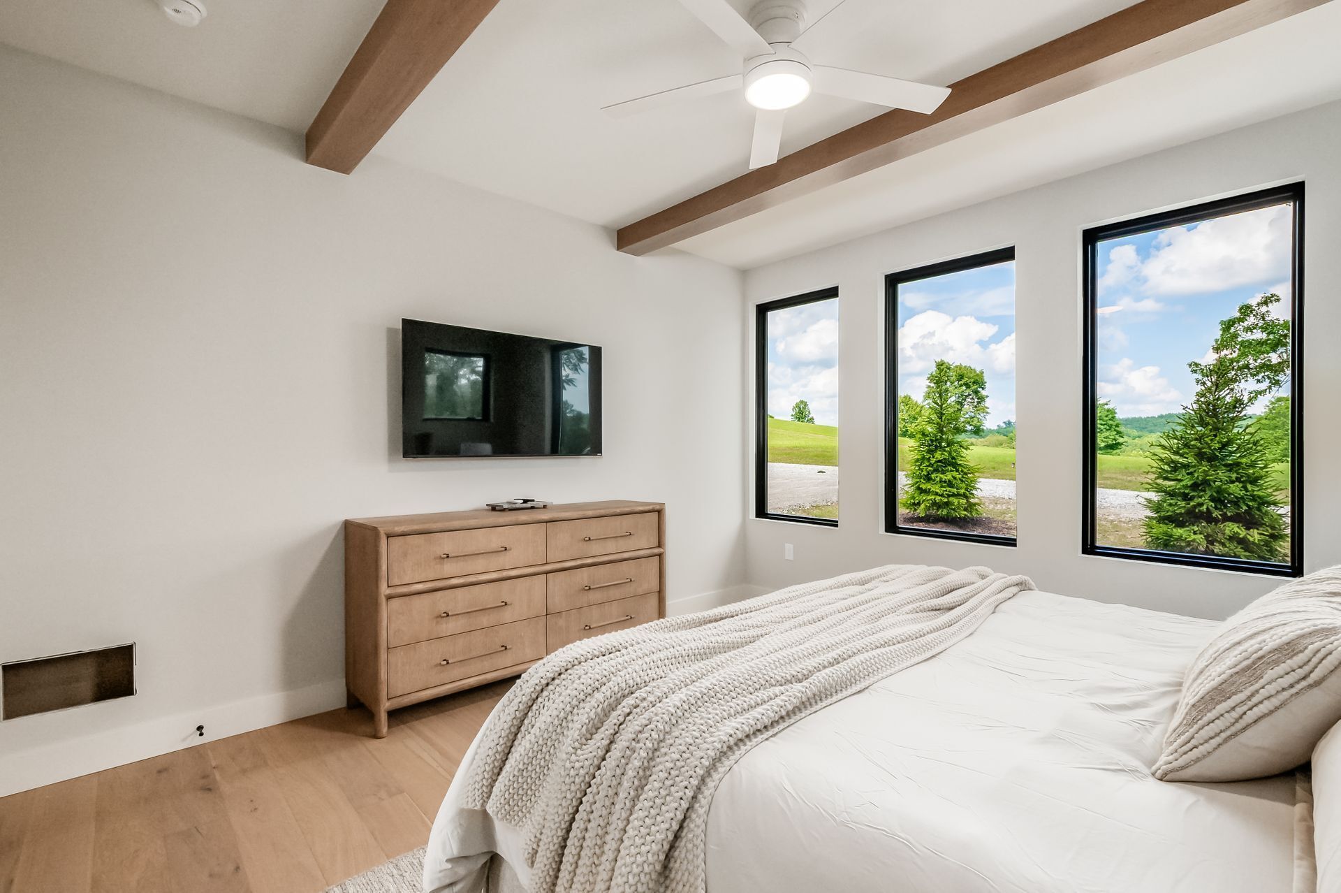 Un dormitorio con cama, cómoda de madera, televisor y ventanas amplias con vistas a follaje.