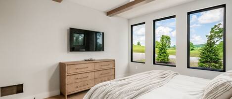 Un dormitorio con cama, cómoda de madera, televisor y ventanas amplias con vistas a follaje.