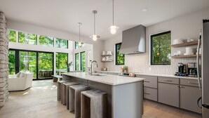 A modern kitchen with a large island, built-in appliances, and large windows offering views of greenery. - Book this Luxury Hocking Hills Lodge! Sleeps 14 (4BR/2.5BA). Enjoy a spa-like bath, full kitchen, fireplace & deck in Arcadia Hills. The ultimate family getaway! (New Plymouth)