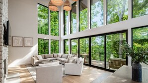 A spacious living area with a large window overlooking a forest, a modern sofa set, and a wooden floor. - Book this Luxury Hocking Hills Lodge! Sleeps 14 (4BR/2.5BA). Enjoy a spa-like bath, full kitchen, fireplace & deck in Arcadia Hills. The ultimate family getaway! (New Plymouth)