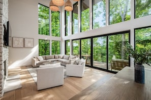 Amplia sala de estar con un ventanal que da a un bosque, un conjunto de sofás modernos y piso de madera.