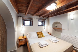 Un dormitorio con techo de madera, dos ventanas con cortinas, una mesita de noche con lámpara, una cama con ropa blanca y un estante pequeño con una planta.