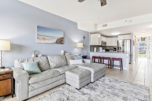 A living room with a sectional sofa, a coffee table, a rug, and a kitchen area with a refrigerator, microwave, and bar stools. - Bay Views & Breezes  ~ Beach & Bay! (Pensacola Beach)