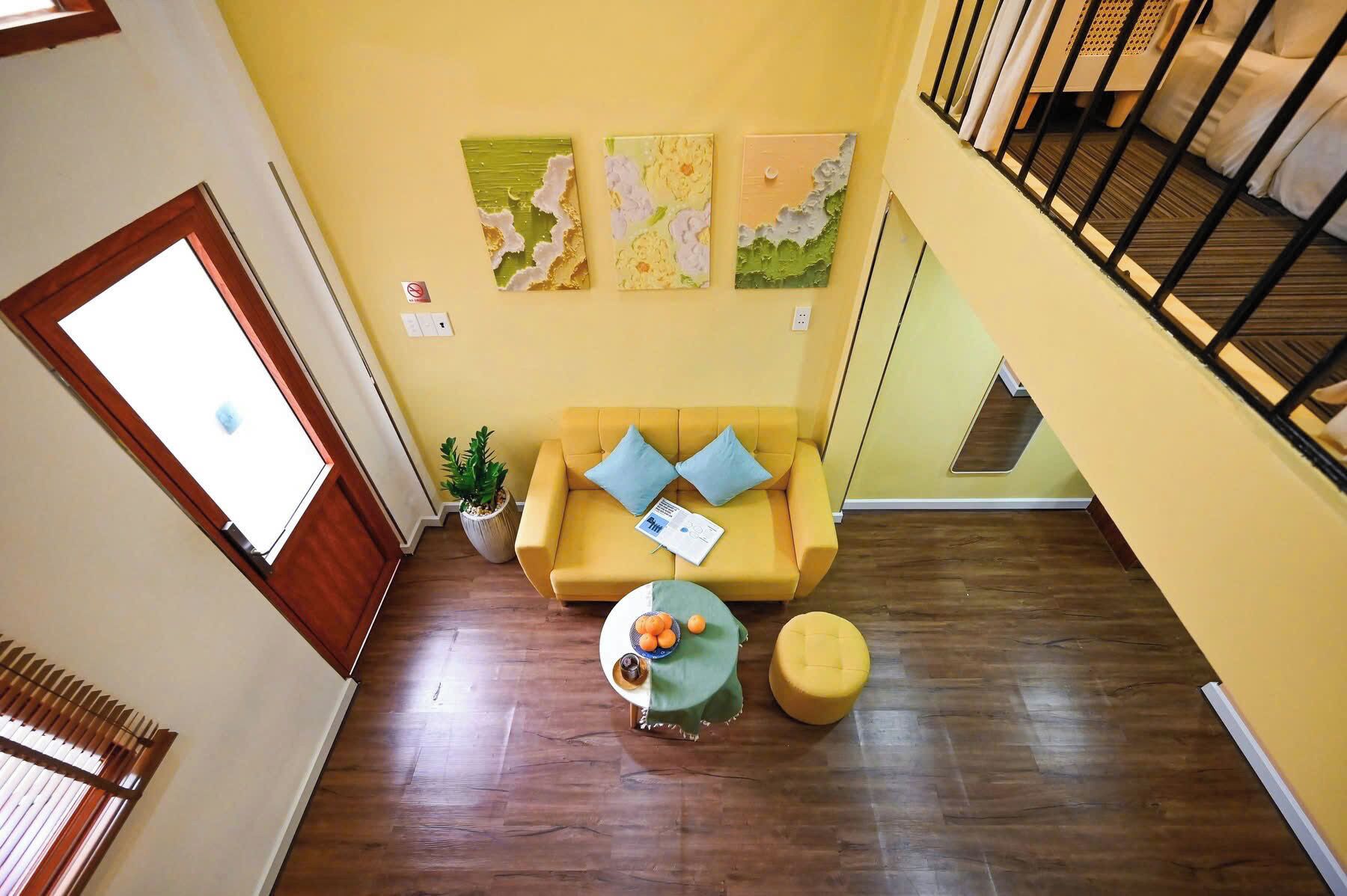 Une pièce avec un canapé jaune, une petite table sur laquelle sont posées des oranges, une plante en pot et trois œuvres d’art abstraites accrochées au mur.