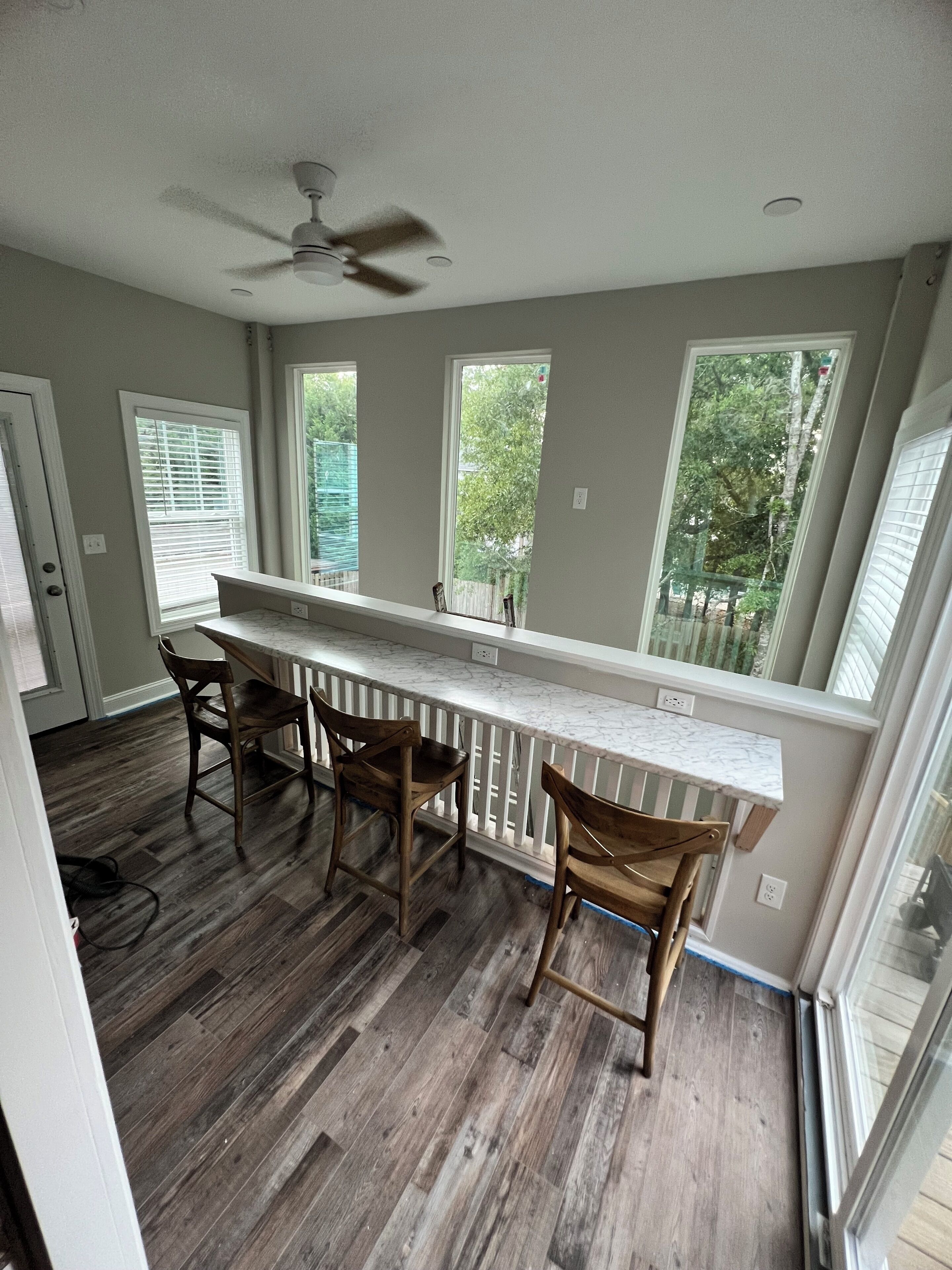 A kitchen with a bar area, wooden chairs, a ceiling fan, and large windows.