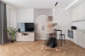 Uma sala de estar moderna com televisão de tela plana, área de jantar e cozinha com armários brancos e detalhes em mosaico estilo herringbone.