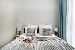 A neatly made bed with a grey striped blanket, two pillows with a cityscape logo, a bedside table with a lamp, and a window with blue curtains. - Towarowa Square & Parking by Downtown Apartments (Warszawa)