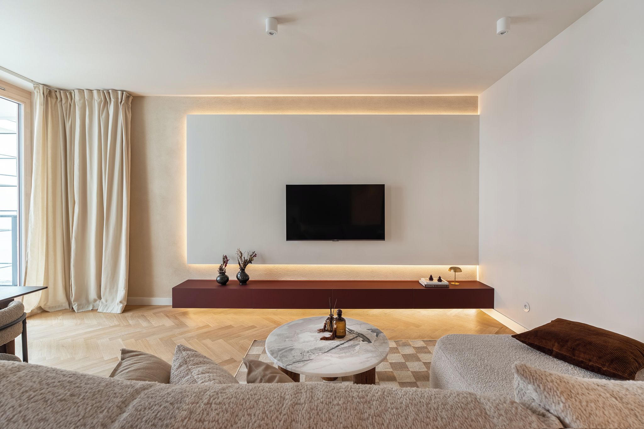 A modern living room with a flat-screen TV mounted on a wall, a dark-colored entertainment unit below, and a round coffee table with decorative items.