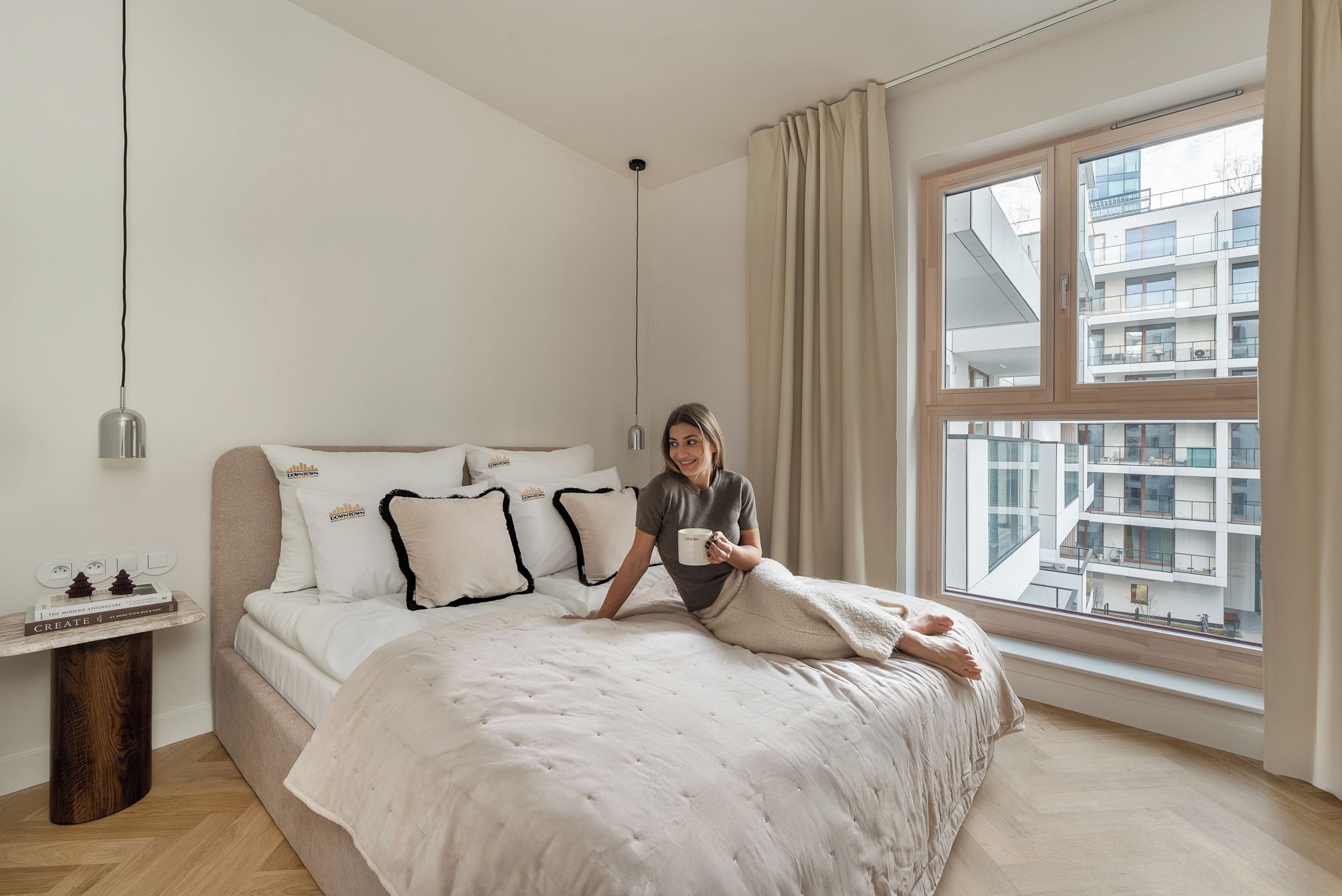 Quarto com cama grande, mesa de cabeceira com livros, janela com vista para edifícios e luminária pendente.