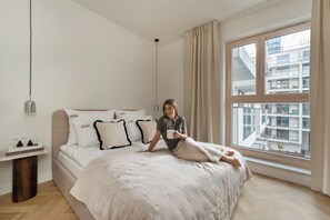 Quarto com cama grande, mesa de cabeceira com livros, janela com vista para edifícios e luminária pendente.