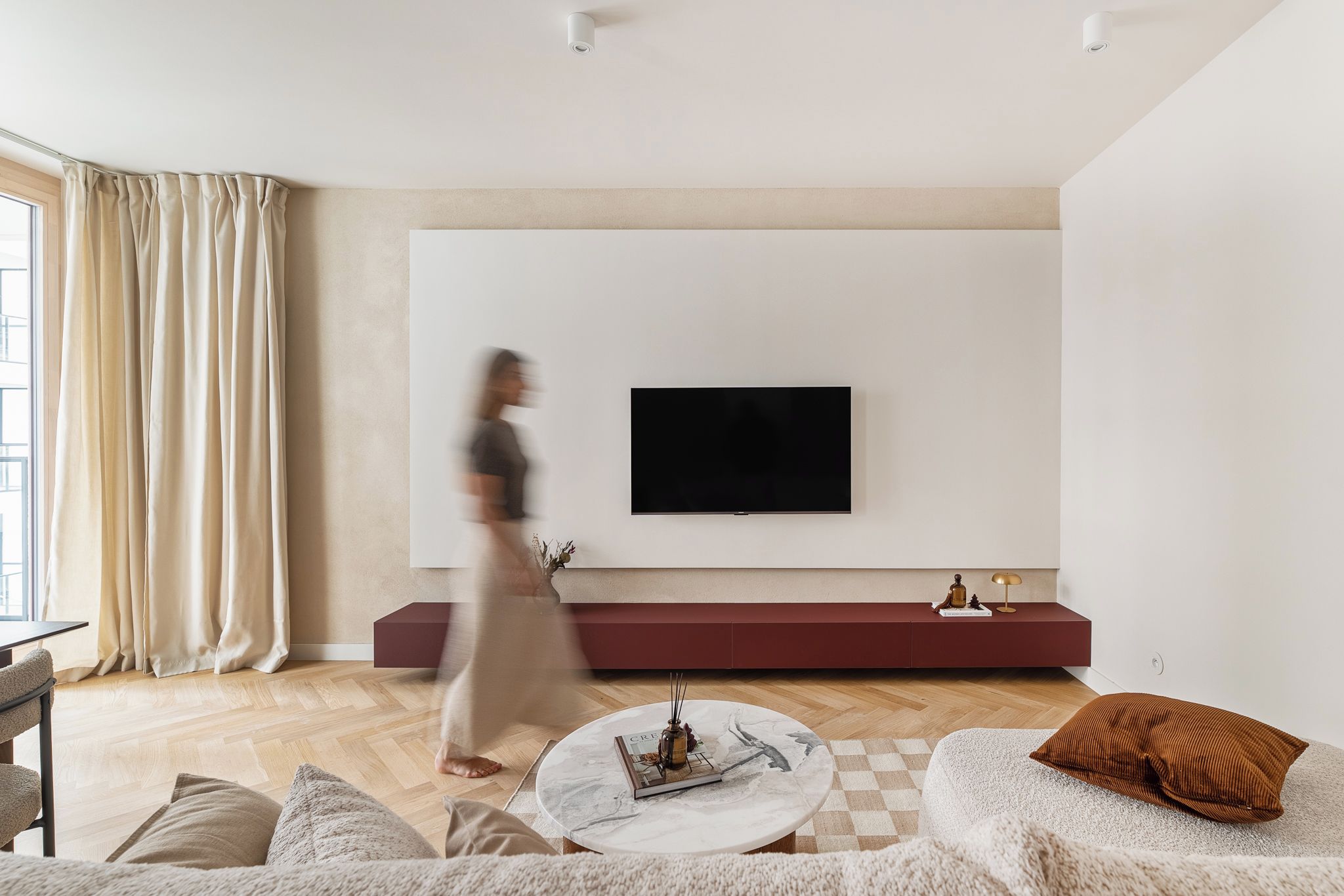 Uma sala de estar moderna com uma televisão de tela plana na parede, uma mesa de centro redonda com um livro e um vaso, um sofá com um travesseiro e um banco vermelho.