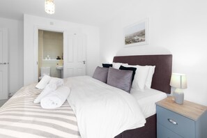 Een net opgemaakt bed met witte lakens, een bedlampje en een klein dressoir.