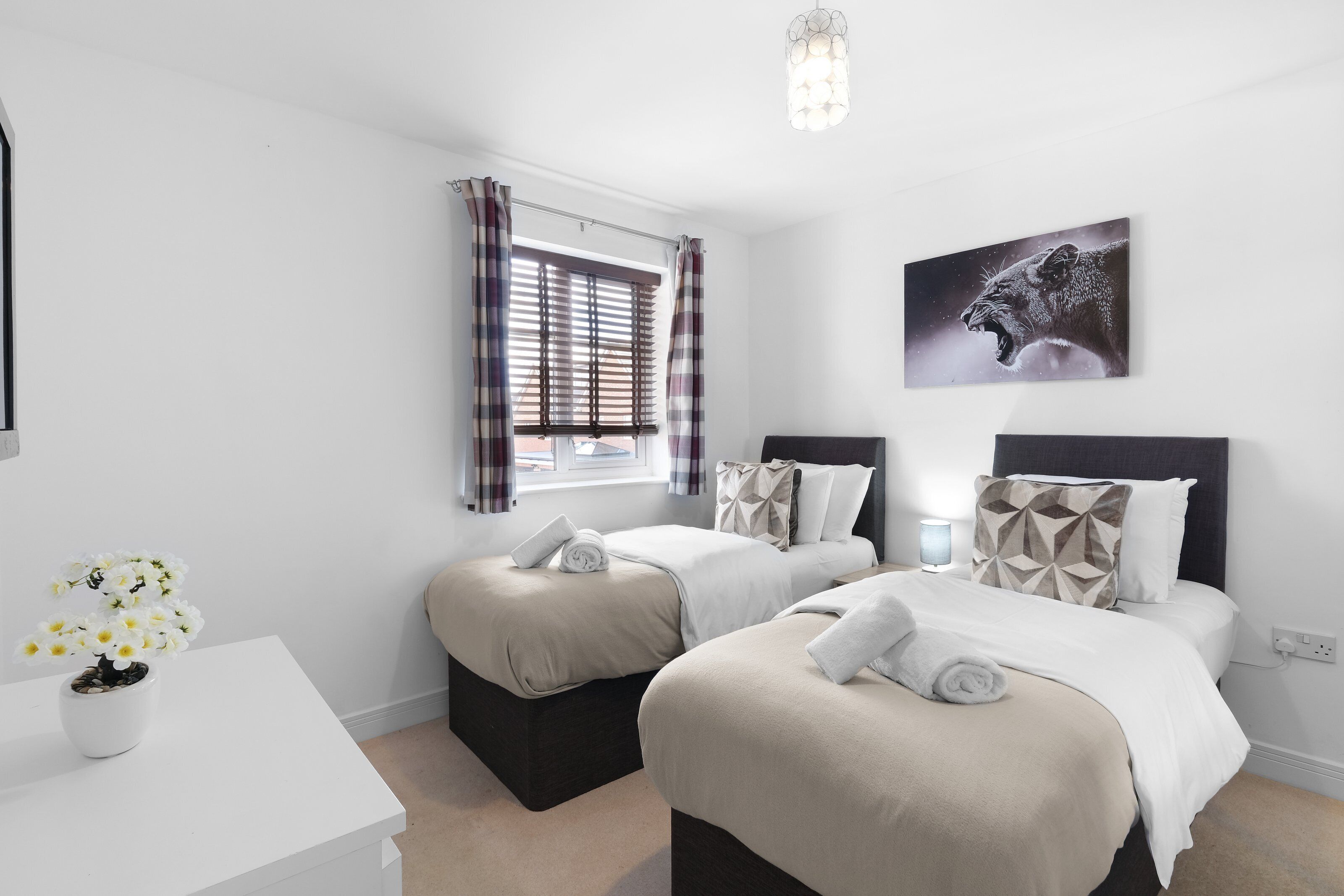 A hotel room with two beds, a window with curtains, a wall-mounted artwork, and a bedside table with a vase of flowers.