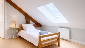 Ein Schlafzimmer mit schrägen Wänden, einem Oberlicht, einem hölzernen Bett, einem Nachttisch mit Lampe und einer weißen Wand.