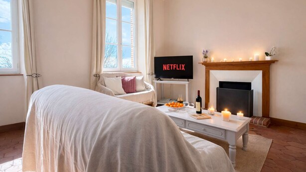 Ein gemütliches Zimmer mit einem Bett, einem Fernseher, der 'NETFLIX' zeigt, einem Kamin sowie einem Tisch mit Kerzen und einer Flasche.