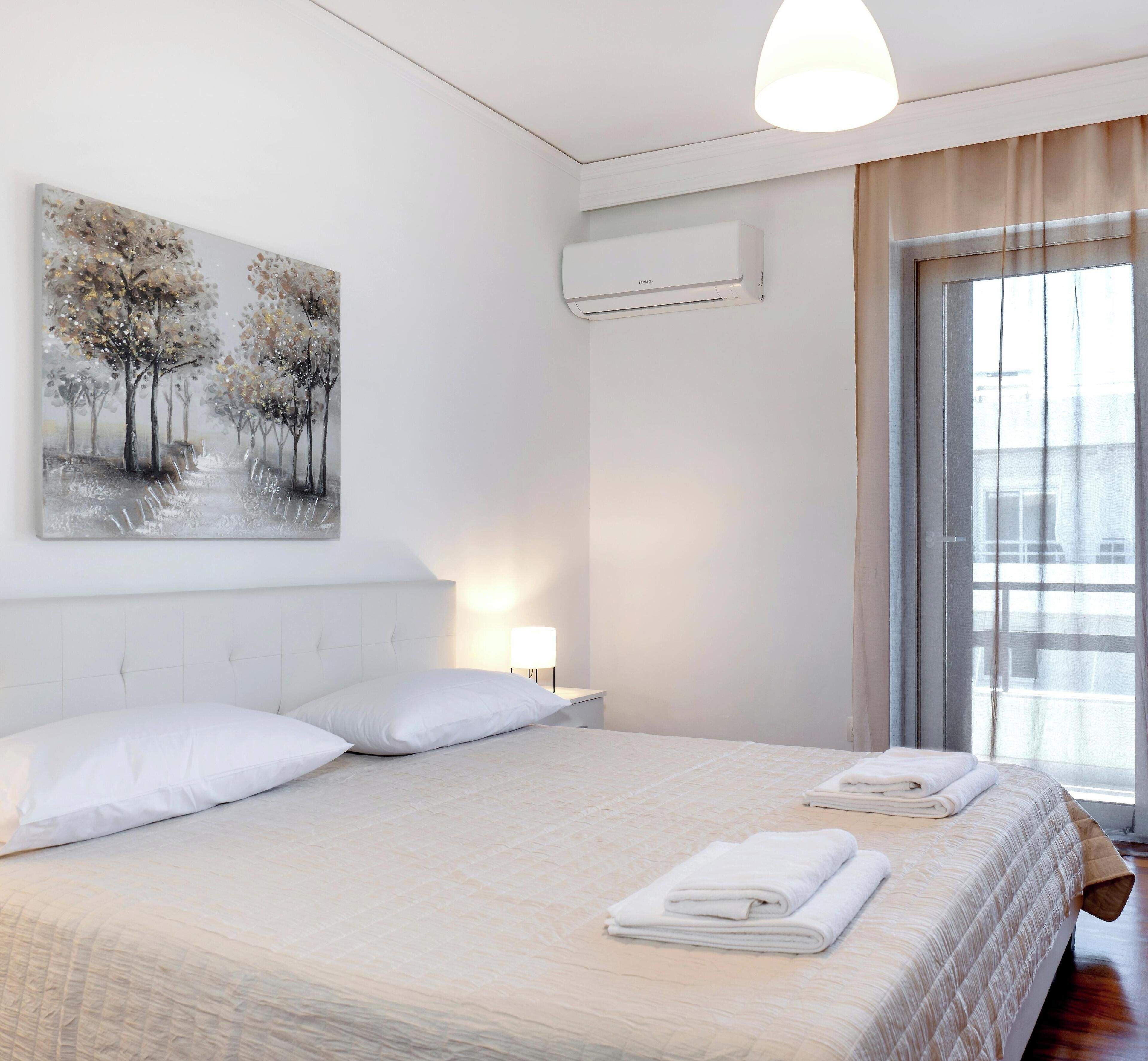 A hotel room with a large bed, white bedding, a painting of trees, an air conditioning unit, and a window with curtains.