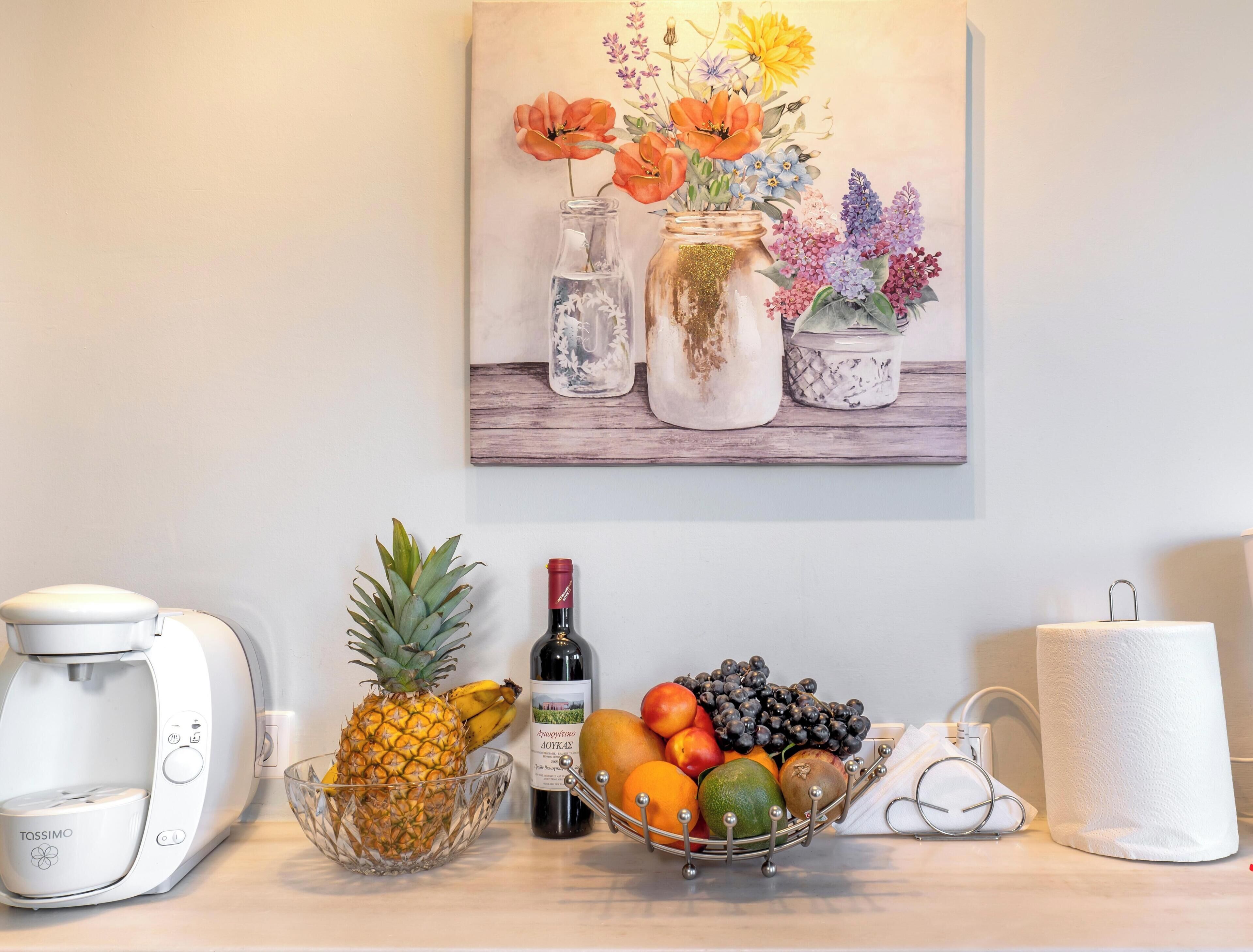 A kitchen counter with a coffee maker, a bowl of fruit, a bottle of wine, and a painting of flowers on the wall.