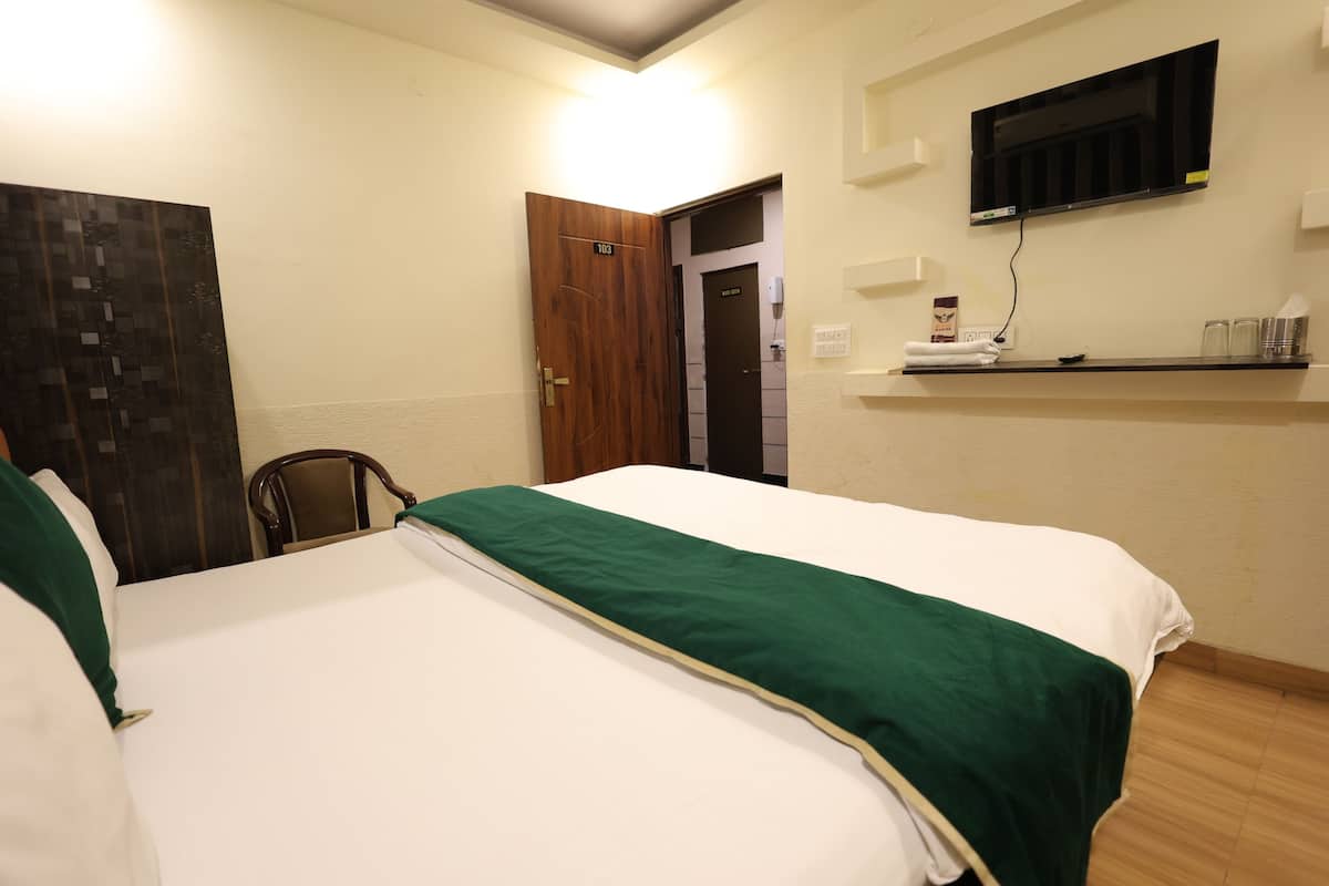 A hotel room with a bed, a wooden door, a TV mounted on the wall, and a small shelf with toiletries.