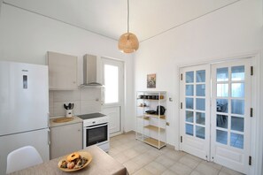 Eine kleine Küche mit Herd, Backofen und weißen Schränken. Ein Esstisch mit einer Fruchtschale und einem gewebten Korb. Eine Korbtreille-Pendelleuchte hängt von der Decke.