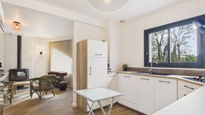 Moderni keittiö, jossa on sisäänrakennettu liesi, ruokailutila pöydän ja tuolien kera sekä suuri ikkuna, josta avautuu näkymä puita kohden.