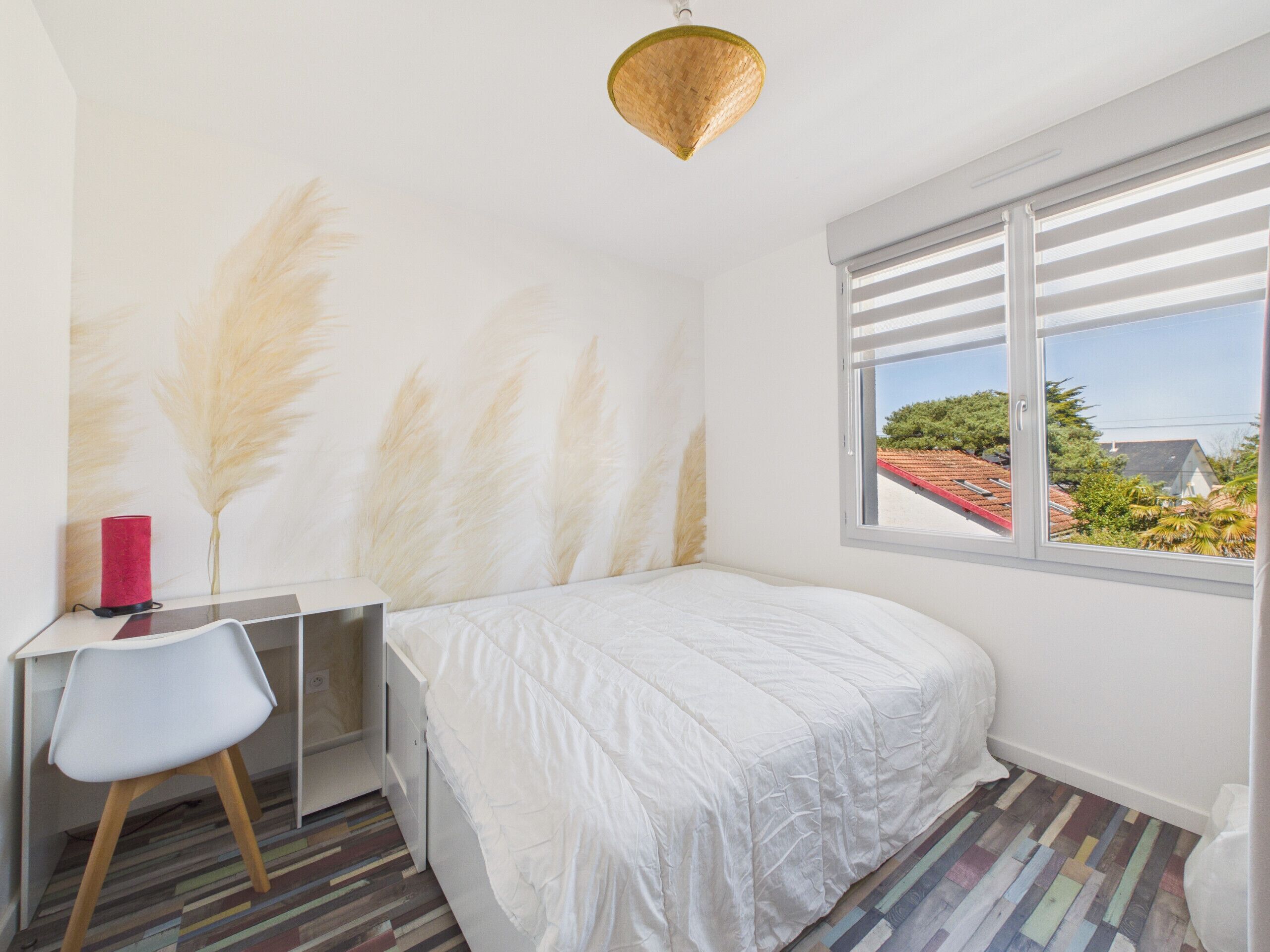 A bedroom with a bed, a desk, a chair, a window with blinds, and a view of the outside.