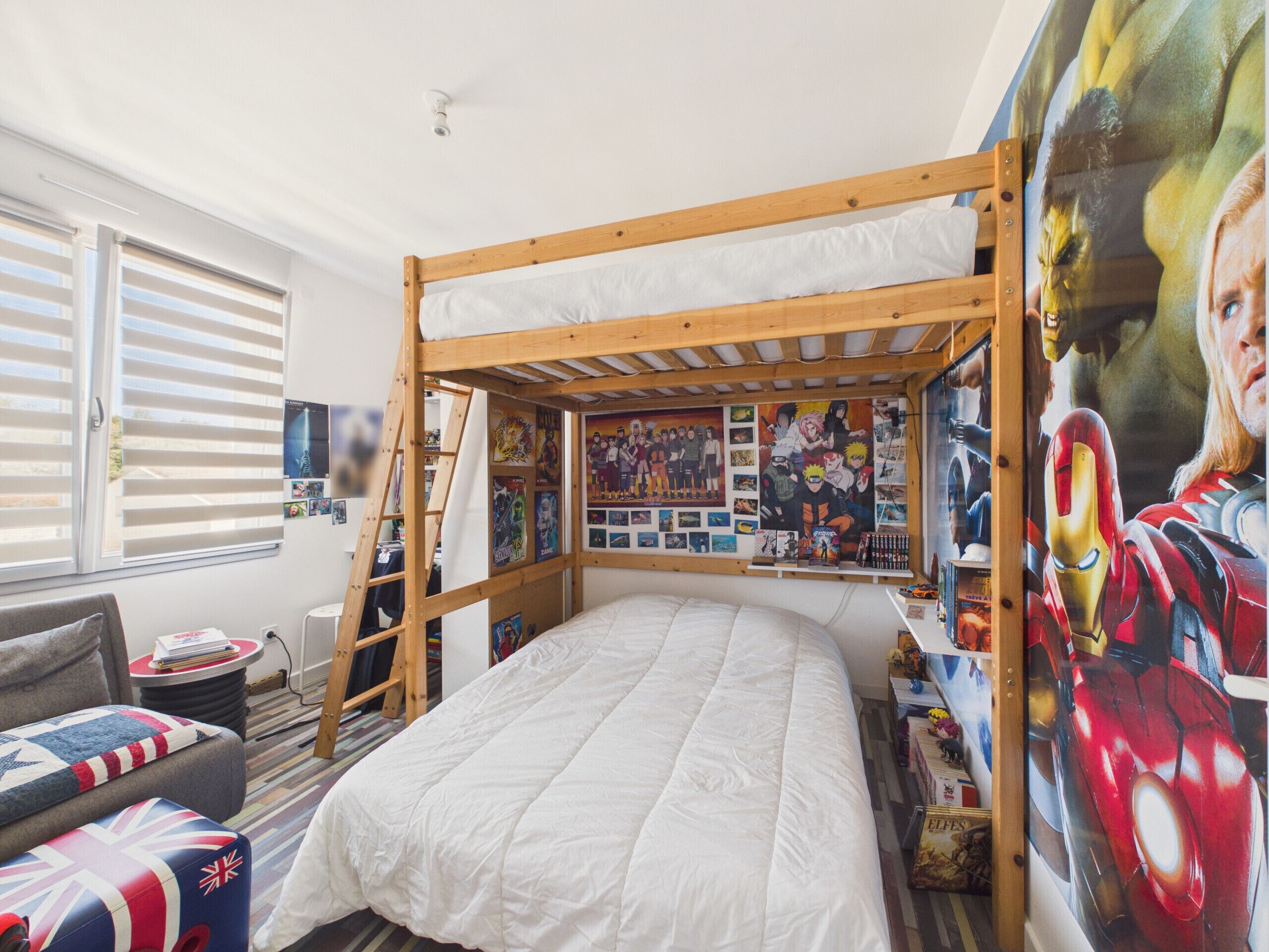 A bunk bed with a wall mural, a window with blinds, a small table, and a couch with a flag cushion.