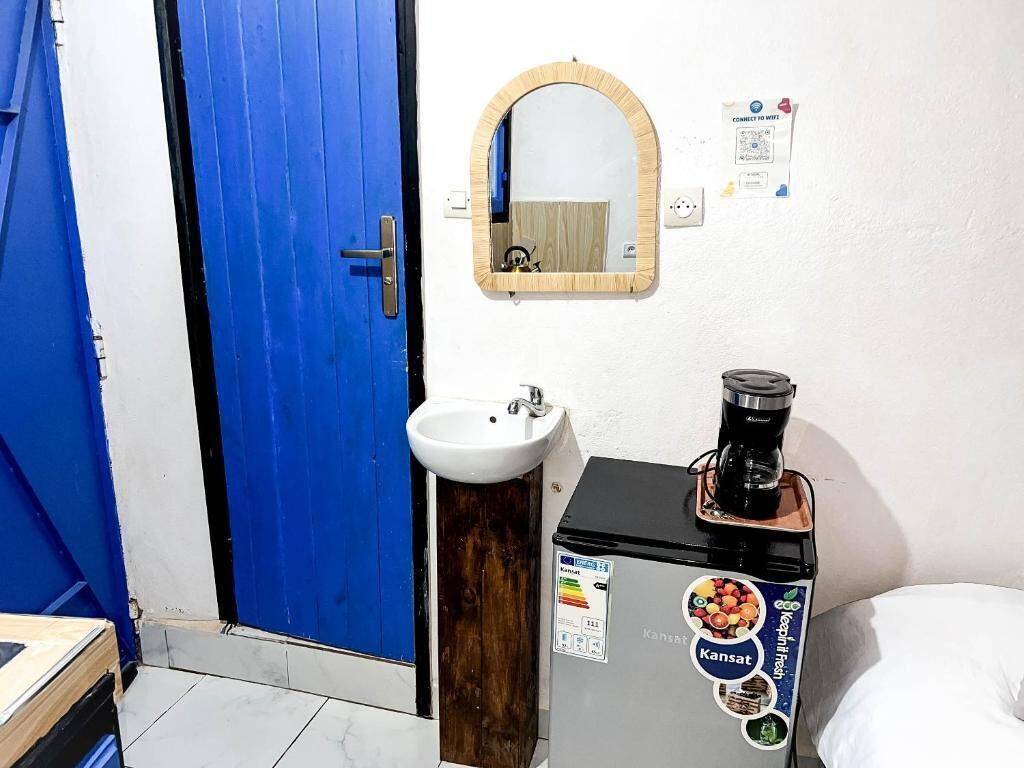 Ein Zimmer mit blauer Tür, Waschbecken in Weiß, Spiegel und Kaffeemaschine auf einem Schrank.
