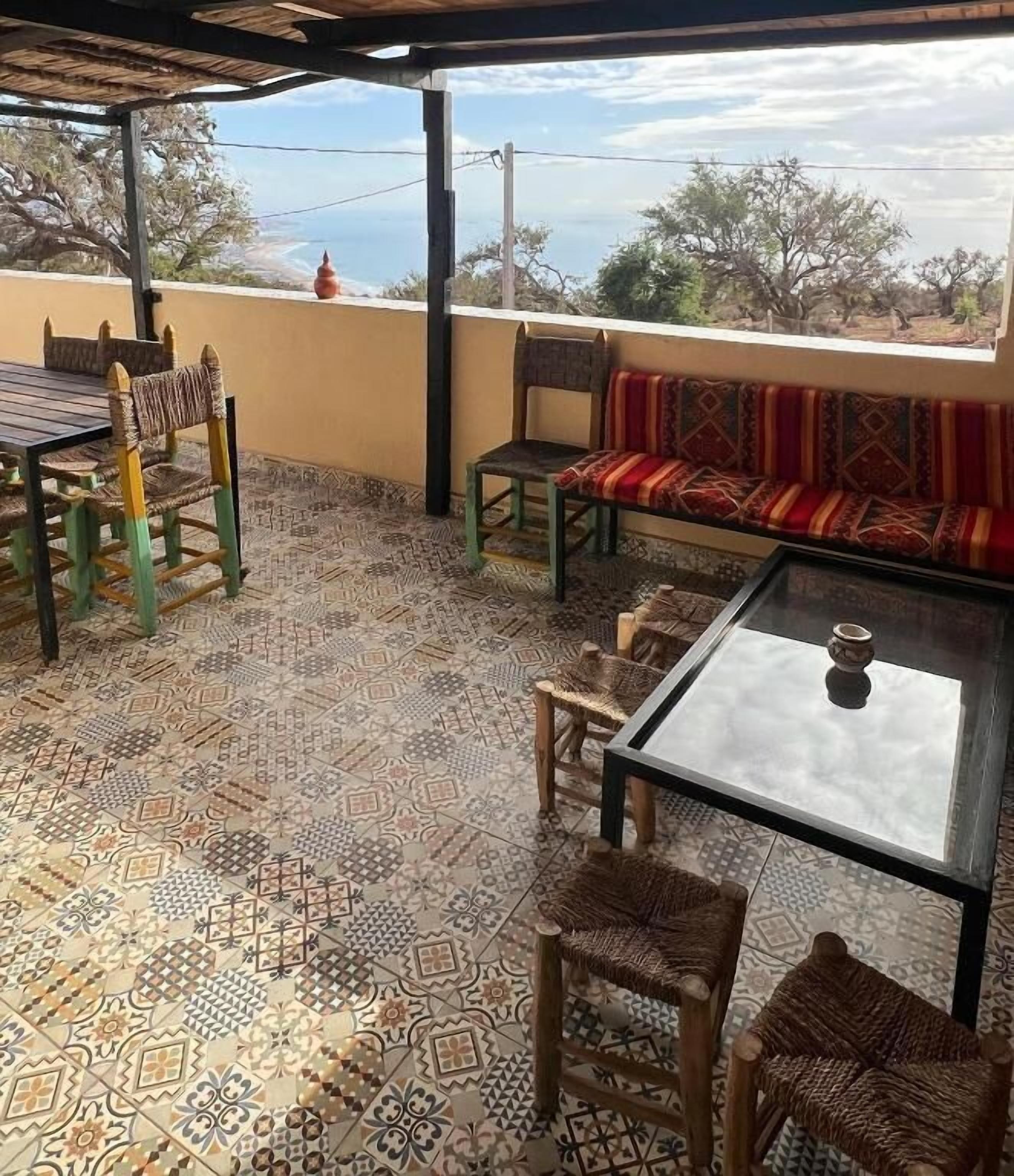 A covered patio with patterned floor tiles, a glass table, wicker chairs, a cushioned bench, and a view of the landscape.
