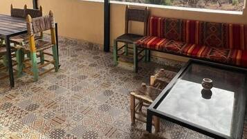A covered patio with patterned floor tiles, a glass table, wicker chairs, a cushioned bench, and a view of the landscape.