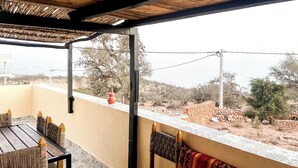 An outdoor patio with a wooden table and chairs, a patterned wall, and a view of a dry landscape with trees and a distant mountain. - Superbo Stay (Taghazout)