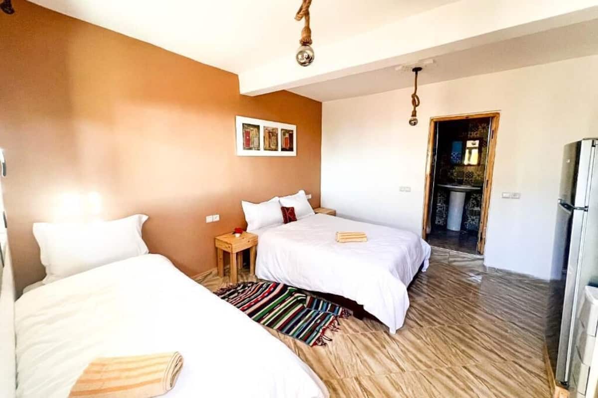 Habitación de hotel con dos camas, piso de madera, refrigerador y puerta que da a un baño.