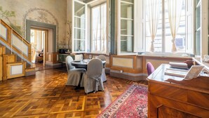 A room with a wooden parquet floor, a dining table with chairs, a desk with a chair, and large windows with curtains.